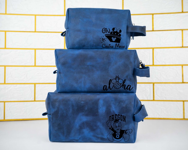 May include: Three blue toiletry bags stacked on top of each other. The top bag has the text "On Cruise Mode" with a cruise ship and Mickey Mouse ears. The middle bag has the text "Aloha" with a pineapple and Mickey Mouse ears. The bottom bag has the text "Oregon" with a cartoon duck wearing a football jersey.