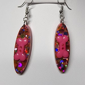 May include: Pink oval shaped earrings with a pink bone charm and iridescent glitter embedded in the resin.