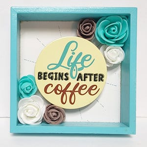 Könnte beinhalten: Ein quadratischer Rahmen in Türkisblau mit weißem Hintergrund und einem gelben Kreis mit dem Text "Life Begins After Coffee" in schwarzer und brauner Schrift. Der Rahmen ist mit weißen, braunen und türkisblauen Stoffblumen verziert.