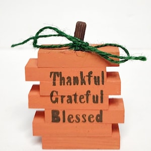 Op de afbeelding: Een kleine houten pompoen met een groene touw strik en een bruine houten steel. De pompoen is oranje geverfd en heeft de woorden "Thankful", "Grateful" en "Blessed" erop geschreven.
