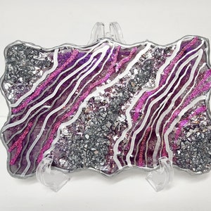 May include: A silver and pink resin coaster with a wavy, abstract design. The coaster has a glittery finish and is set on a clear stand.