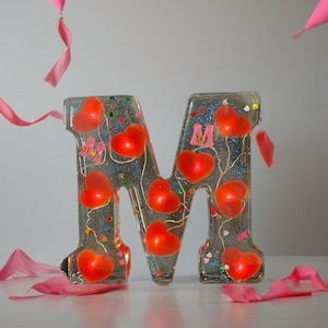 Puede incluir: Una letra "M" de resina transparente llena de detalles en forma de corazón rojo y purpurina. La letra está decorada con pequeños corazones rosas y rodeada de cintas rosas y globos rosas flotantes. Un artículo decorativo.