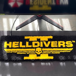 Puede incluir: Alfombra rectangular negra con el texto amarillo "HELLDIVERS" y una calavera con alas. La alfombra está colocada sobre una superficie oscura, con un monitor de ordenador al fondo.