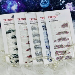 May include: Four packs of nail accessories are displayed, each with a different color scheme of rhinestones and glitter. The packaging reads "TRENDY NAILS ACCESSORIES". A string of pearls is draped in front of the packs, against a fluffy white background.