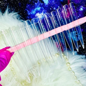 May include: A set of clear plastic nail tips arranged on a pink holder. The tips are long and pointed, and they are designed for use with nail extensions.