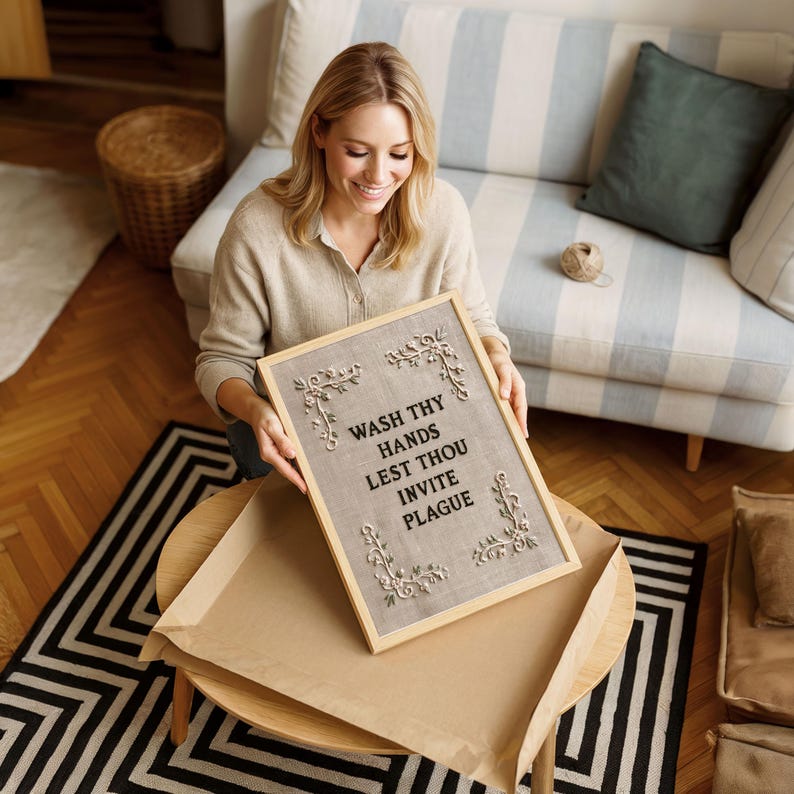 May include: A rectangular wooden framed sign featuring a light brown fabric background with the black text WASH THY HANDS LEST THOU INVITE PLAGUE. The sign is decorated with four ornate, light-colored floral corner accents in a relief style.