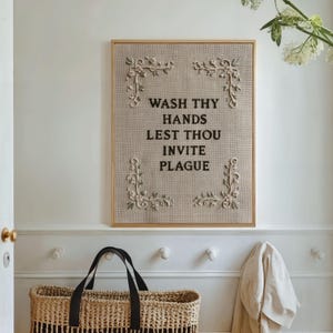May include: A rectangular wall sign featuring a light brown textured fabric background with black embroidered text that reads WASH THY HANDS LEST THOU INVITE PLAGUE. The sign is framed in light wood and decorated with white floral embroidery in each corner.