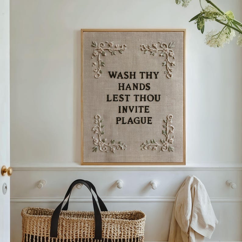 May include: A rectangular wall sign featuring a light brown textured fabric background with black embroidered text that reads WASH THY HANDS LEST THOU INVITE PLAGUE. The sign is framed in light wood and decorated with white floral embroidery in each corner.