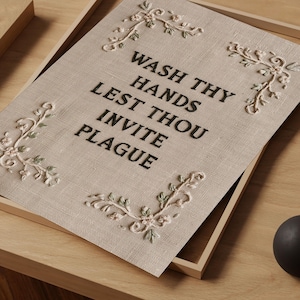 May include: A rectangular beige fabric sign featuring black embroidered text that reads WASH THY HANDS LEST THOU INVITE PLAGUE. The sign is decorated with four embroidered floral corner accents in shades of light pink and sage green.