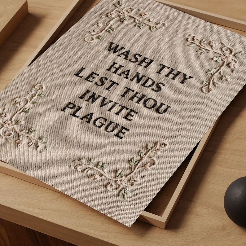 May include: A rectangular beige fabric sign featuring black embroidered text that reads WASH THY HANDS LEST THOU INVITE PLAGUE. The sign is decorated with four embroidered floral corner accents in shades of light pink and sage green.