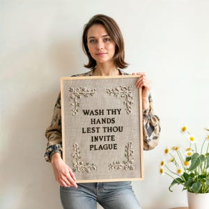 May include: A rectangular wall sign featuring a light brown linen background with black embroidered text that reads WASH THY HANDS LEST THOU INVITE PLAGUE. The sign is framed in light wood and decorated with white floral embroidery in each corner.