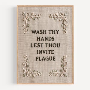 May include: A rectangular wall art piece featuring a light beige linen fabric background framed in light wood. The text WASH THY HANDS LEST THOU INVITE PLAGUE is embroidered in black thread. Four floral corner motifs in cream and green accent the text.