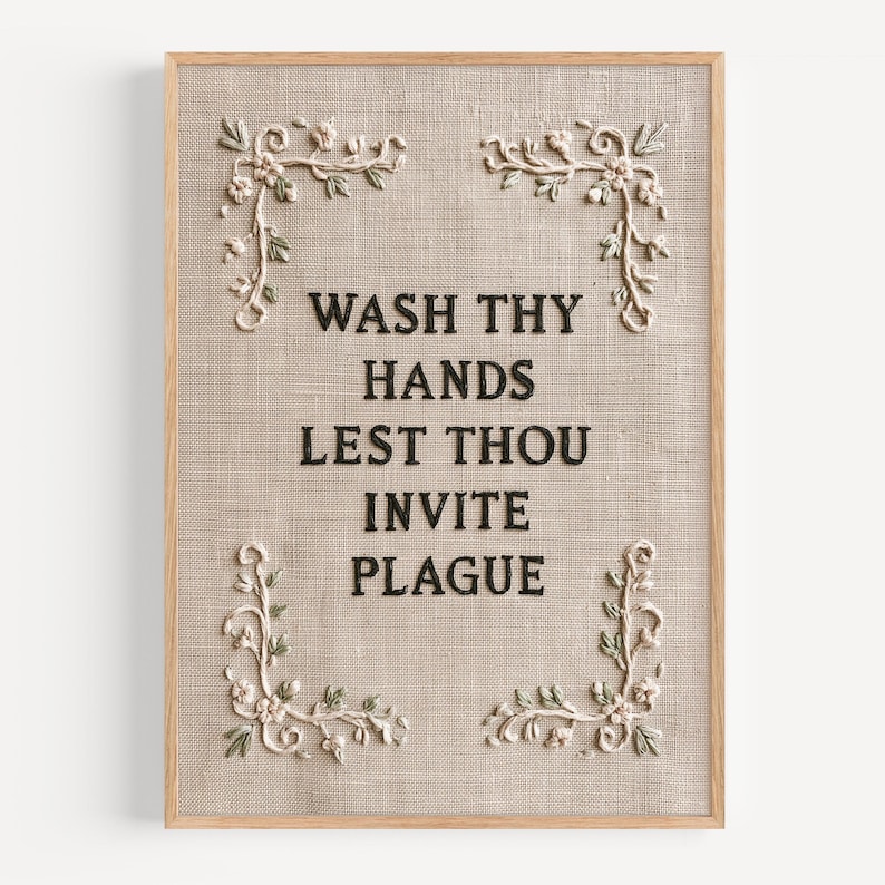 May include: A rectangular wall art piece featuring a light beige linen fabric background framed in light wood. The text WASH THY HANDS LEST THOU INVITE PLAGUE is embroidered in black thread. Four floral corner motifs in cream and green accent the text.