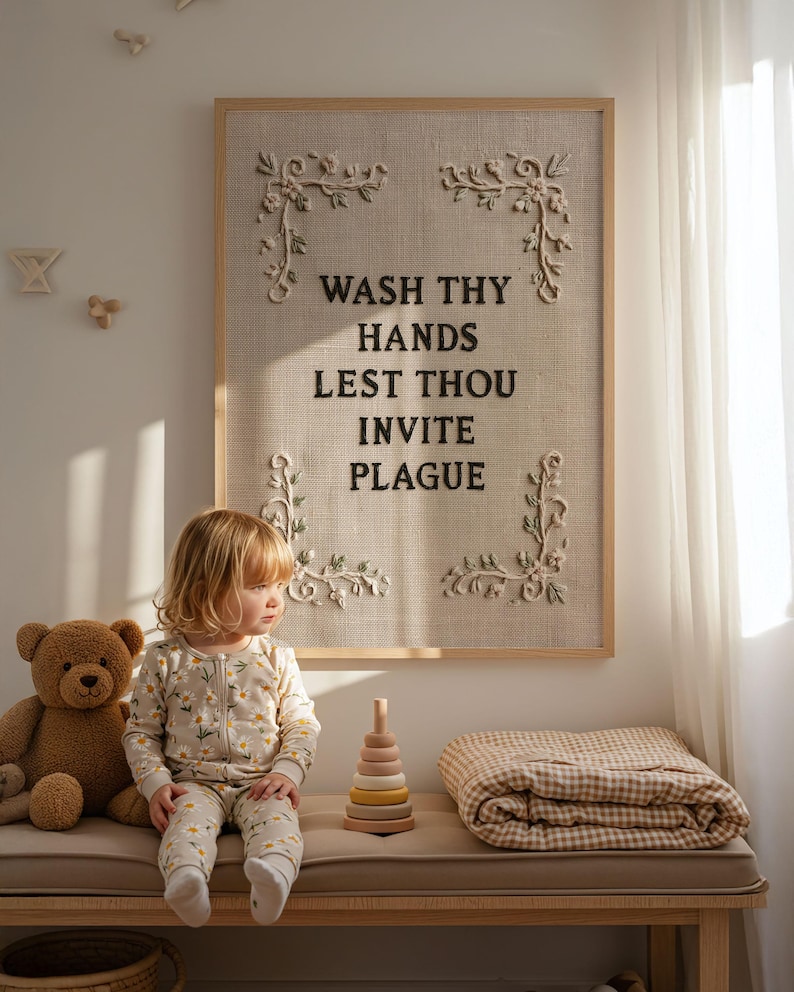 May include: A rectangular wall art piece featuring a light beige textured fabric background framed in light wood. The text WASH THY HANDS LEST THOU INVITE PLAGUE is embroidered in dark brown thread, accented by four cream-colored floral corner motifs.