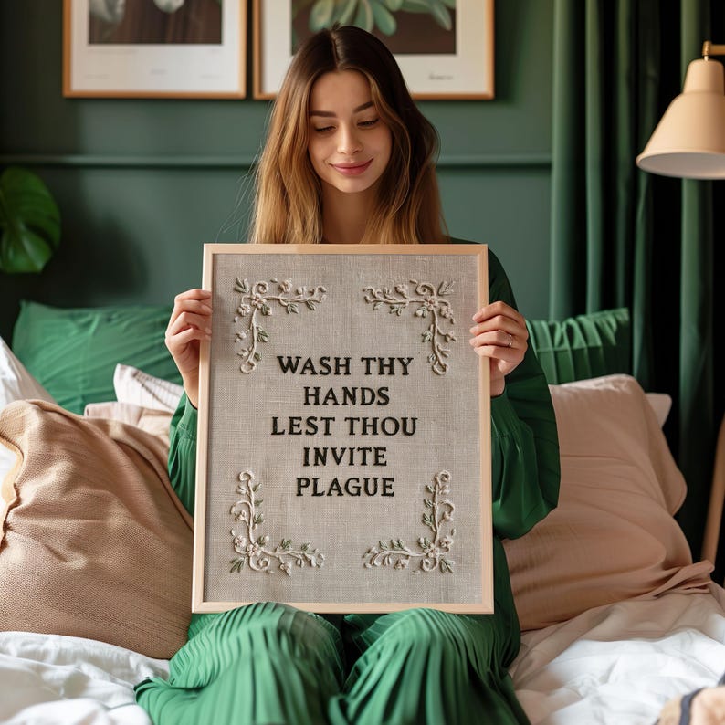 May include: A rectangular framed wall art piece featuring a light gray fabric background with black embroidered text that reads WASH THY HANDS LEST THOU INVITE PLAGUE. The design includes four decorative white floral corner accents.