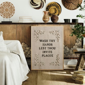 May include: A rectangular framed wall art piece featuring a light beige fabric background with the black text WASH THY HANDS LEST THOU INVITE PLAGUE. The design includes four white floral vine corner embellishments on a textured linen-like surface.