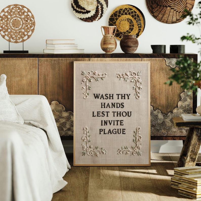 May include: A rectangular framed wall art piece featuring a light beige fabric background with the black text WASH THY HANDS LEST THOU INVITE PLAGUE. The design includes four white floral vine corner embellishments on a textured linen-like surface.