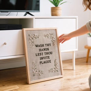 May include: A rectangular framed wall art piece featuring a light beige linen background with black embroidered text that reads WASH THY HANDS LEST THOU INVITE PLAGUE. The design includes delicate white floral embroidery in each of the four corners.