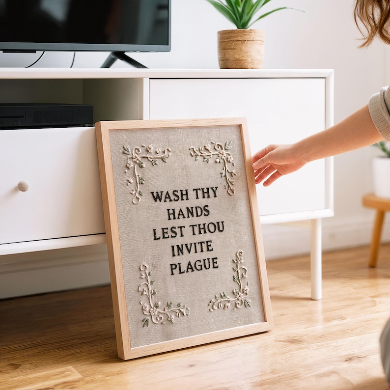 May include: A rectangular framed wall art piece featuring a light beige linen background with black embroidered text that reads WASH THY HANDS LEST THOU INVITE PLAGUE. The design includes delicate white floral embroidery in each of the four corners.