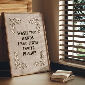 May include: A rectangular framed wall art piece featuring a beige textured fabric background. The text WASH THY HANDS LEST THOU INVITE PLAGUE is embroidered in black. Four corners are decorated with white and green floral embroidery motifs.