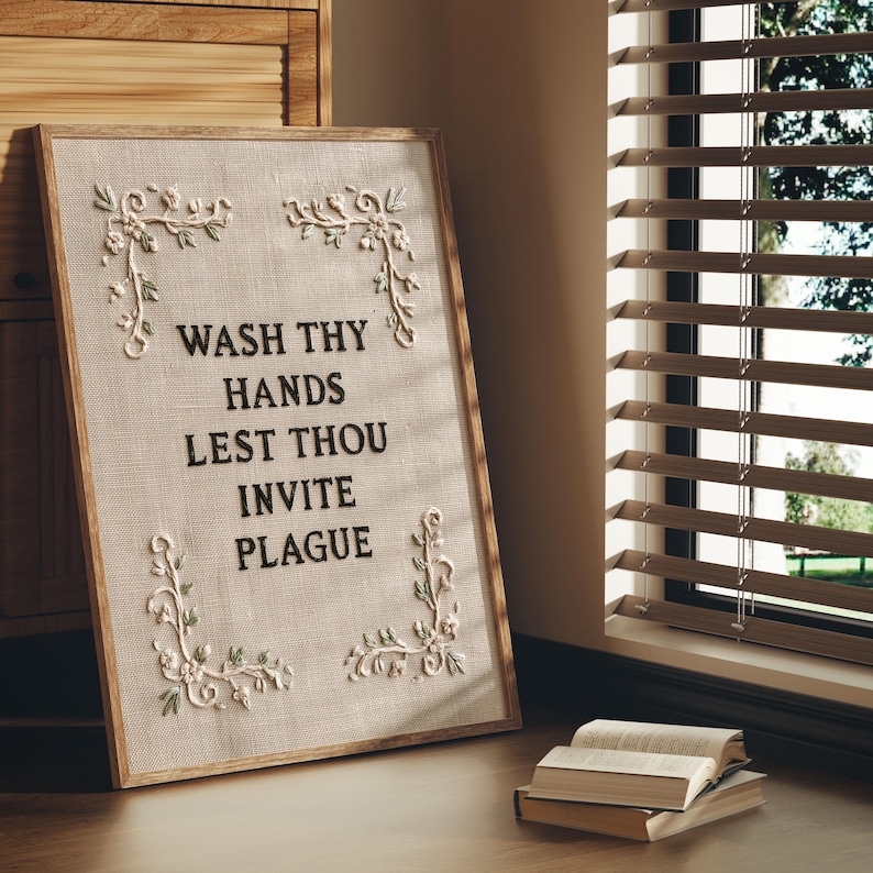May include: A rectangular framed wall art piece featuring a beige textured fabric background. The text WASH THY HANDS LEST THOU INVITE PLAGUE is embroidered in black. Four corners are decorated with white and green floral embroidery motifs.