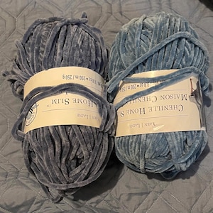 May include: Two skeins of blue chenille yarn. Each skein is wrapped with a paper label that reads "CHENILLE HOME SLIM" and "YARN | LAINE". The yarn is a soft, plush material, perfect for knitting or crocheting projects.