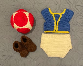 Könnte beinhalten: Ein handgefertigtes Baby-Outfit aus Häkelarbeit mit einem rot-weißen pilzförmigen Hut, einer blauen Weste mit gelben Verzierungen, einer weißen Windel und braunen Babyschuhen. Das Set ist für ein Baby entworfen und aus weichem Garn gefertigt.