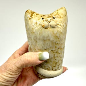 May include: A ceramic cat-shaped mug with a beige and brown speckled glaze. The mug features a detailed cat face with whiskers and a smiling expression. The mug has a rounded base and a tall, slender body.