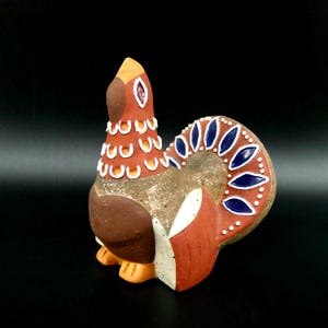 May include: A ceramic bird figurine with a brown body, orange beak and feet, and a decorative tail. The tail features blue and white accents. The bird has a textured, speckled appearance and is set against a black background.