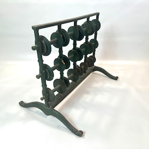 Könnte beinhalten: Ein antiker grüner Metall-Garnrollenhalter. Der Halter hat mehrere Reihen von Spulen, mit einer Vielzahl von Größen. Der Rahmen hat einen dekorativen, gebogenen Sockel und eine rechteckige Oberseite. Die Spulen sind eine Mischung aus Grün und Braun.