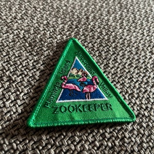 Könnte beinhalten: Ein grüner dreieckiger Aufnäher mit den Worten "Flamingo Island Neverland Valley" und "Zookeeper". Der Aufnäher zeigt ein Design von rosa Flamingos in einem blauen See mit Bergen im Hintergrund.