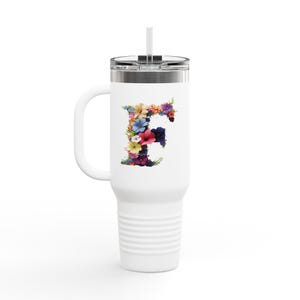 Può includere: Tazza da viaggio bianca con manico e design floreale a forma di "F". La lettera è composta da fiori colorati nei toni del rosa, blu, giallo e viola. La tazza ha un coperchio argentato e una cannuccia.