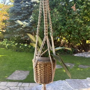 Puede incluir: Un macramé para colgar plantas con una cesta tejida que contiene una planta verde. El macramé está hecho con cordón de color natural y cordón verde. El macramé está colgado de un gancho.