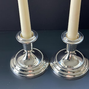 May include: Two silver candle holders with ivory-colored candles. The candle holders have a round base with a beaded edge and a flared stem. The candles are straight and centered in the holders. The background is a dark gray.