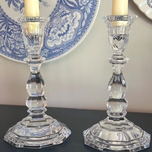 May include: A pair of clear glass candlesticks featuring a faceted, multi-tiered base and a bulbous, rounded stem design. Each candlestick holds a single cream-coloured taper candle within a textured, scalloped top rim, showcasing a reflective finish.