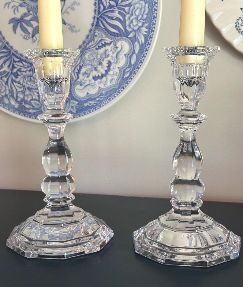 May include: A pair of clear glass candlesticks featuring a faceted, multi-tiered base and a bulbous, rounded stem design. Each candlestick holds a single cream-coloured taper candle within a textured, scalloped top rim, showcasing a reflective finish.