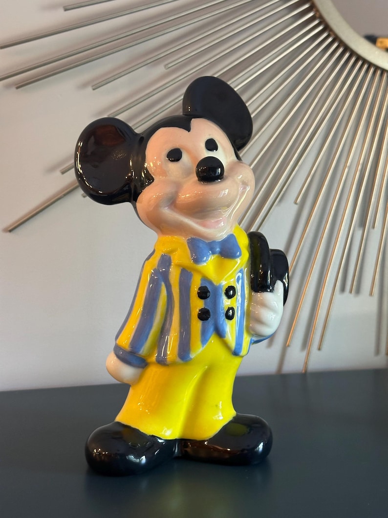 May include: A ceramic figurine of Mickey Mouse wearing a yellow and blue striped suit with a black bow tie. He is holding a black hat in his right hand and has a big smile on his face.