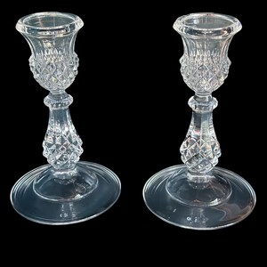May include: A pair of clear glass candle holders featuring a flared top rim, a textured diamond-cut pattern on the upper cup and central stem, and a smooth, wide circular base. The glass is transparent and reflects light across its faceted surface.