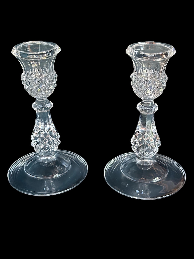 May include: A pair of clear glass candle holders featuring a flared top rim, a textured diamond-cut pattern on the upper cup and central stem, and a smooth, wide circular base. The glass is transparent and reflects light across its faceted surface.