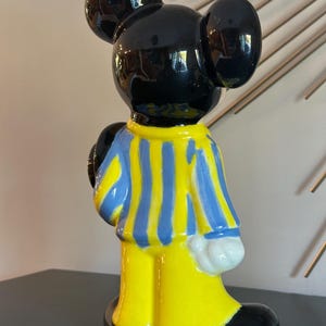 May include: A ceramic figurine of Mickey Mouse wearing a blue and yellow striped shirt and yellow pants. The figurine has black ears and shoes.