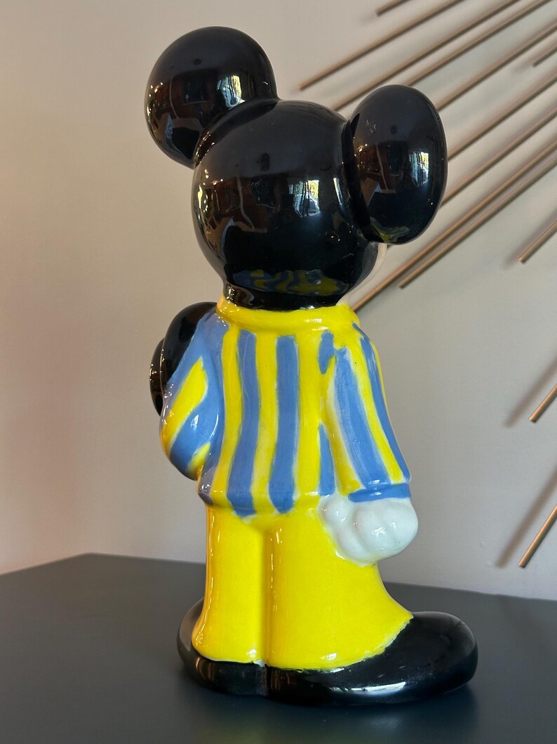 May include: A ceramic figurine of Mickey Mouse wearing a blue and yellow striped shirt and yellow pants. The figurine has black ears and shoes.