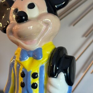 May include: A ceramic figurine of Mickey Mouse wearing a yellow and blue striped jacket with a blue bow tie. He is holding a black top hat in his right hand.