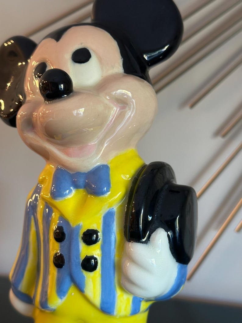 May include: A ceramic figurine of Mickey Mouse wearing a yellow and blue striped jacket with a blue bow tie. He is holding a black top hat in his right hand.
