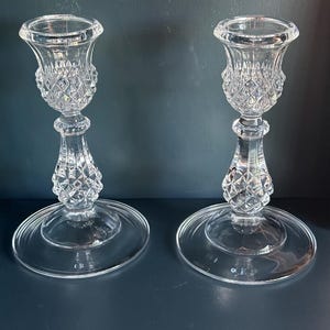 May include: A pair of clear glass candle holders featuring a faceted diamond-cut pattern on the upper cup and the central stem. Each holder has a wide, smooth, circular base and a flared top rim designed to securely hold a standard taper candle.