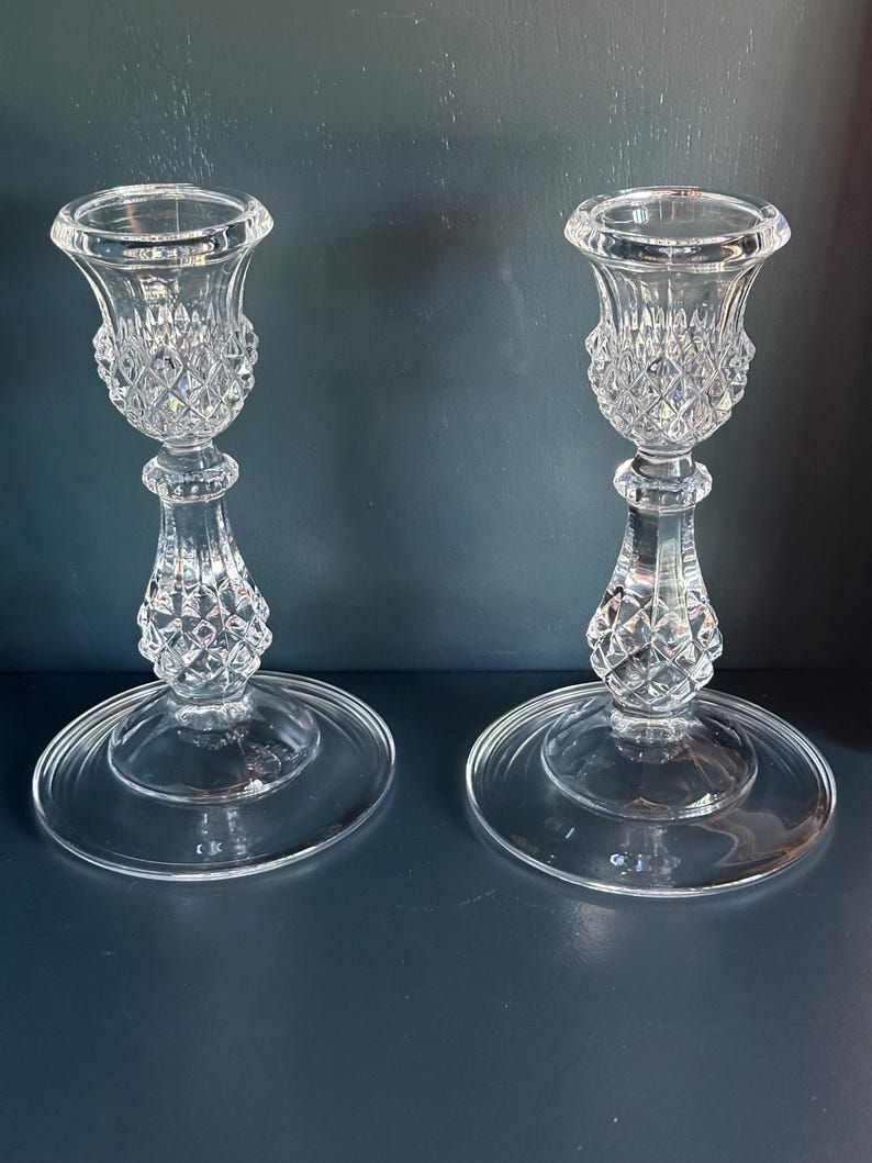 May include: A pair of clear glass candle holders featuring a faceted diamond-cut pattern on the upper cup and the central stem. Each holder has a wide, smooth, circular base and a flared top rim designed to securely hold a standard taper candle.