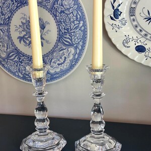 May include: Two clear glass candle holders with a faceted, multi-sided base and a rounded, bulbous stem design. Each holder contains a tall, slender, cream-coloured taper candle. The glass features a reflective, polished surface with clear edges.