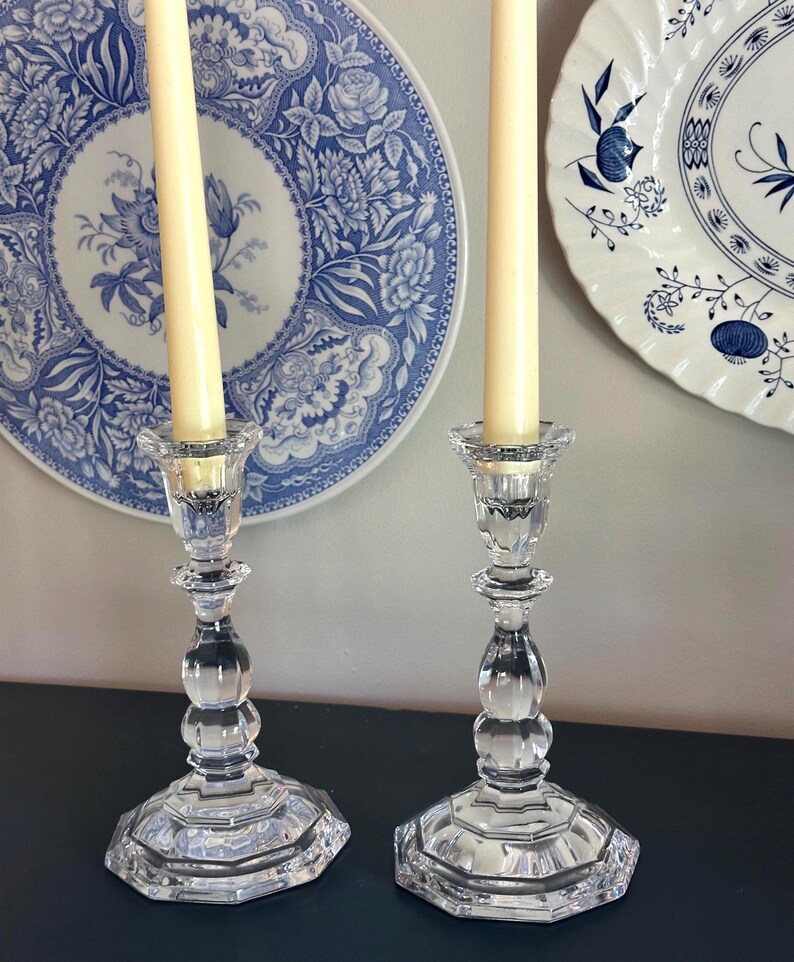 May include: Two clear glass candle holders with a faceted, multi-sided base and a rounded, bulbous stem design. Each holder contains a tall, slender, cream-coloured taper candle. The glass features a reflective, polished surface with clear edges.