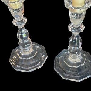 May include: A pair of clear glass candlesticks featuring an octagonal base and a faceted, multi-tiered stem design. Each candlestick holds a single cream-coloured taper candle, showcasing a reflective, transparent surface and geometric structure.