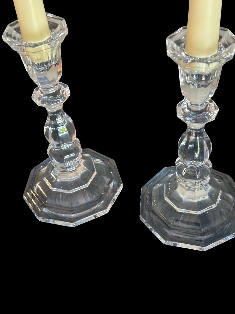 May include: A pair of clear glass candlesticks featuring an octagonal base and a faceted, multi-tiered stem design. Each candlestick holds a single cream-coloured taper candle, showcasing a reflective, transparent surface and geometric structure.