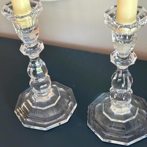 May include: A pair of clear glass candle holders with an octagonal base and a faceted, tiered stem design. Each holder features a scalloped top rim and contains a single, smooth, cream-coloured taper candle inserted into the central socket.